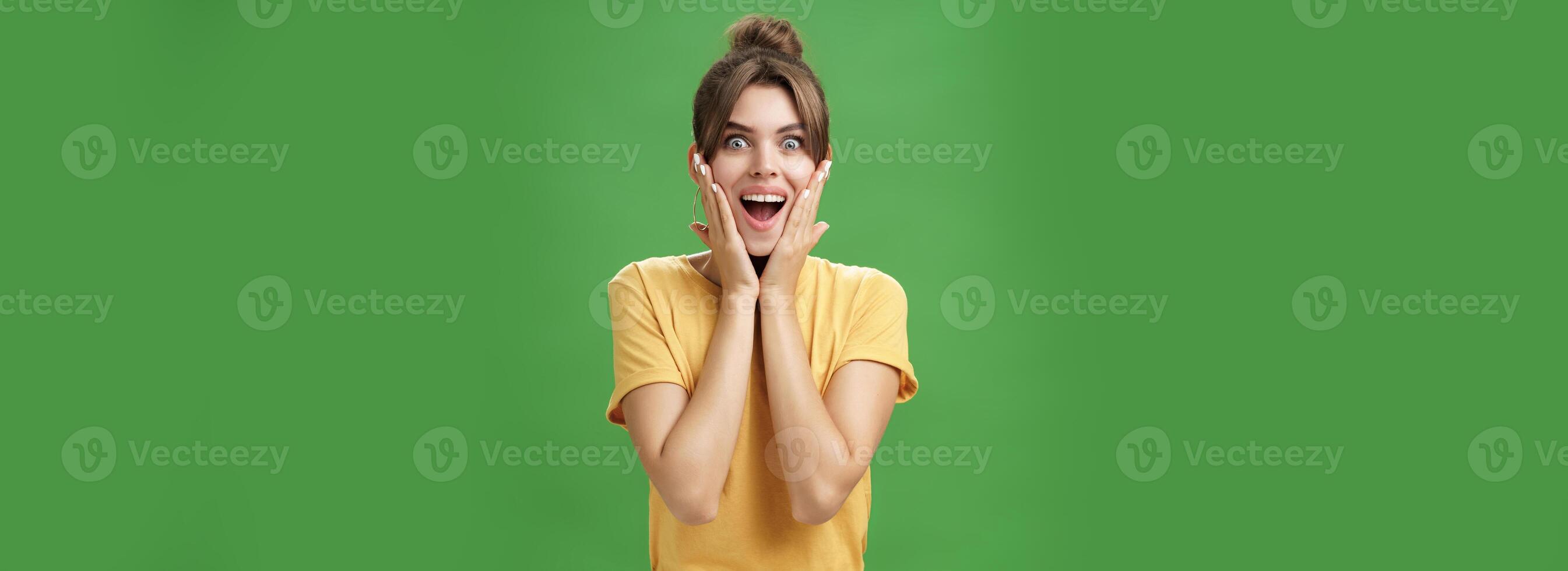 Indoor shot of excited and surprised energized attractive glamorous female in yellow t-shirt touching cheeks from amazement smiling broadly astonished posing over green background photo