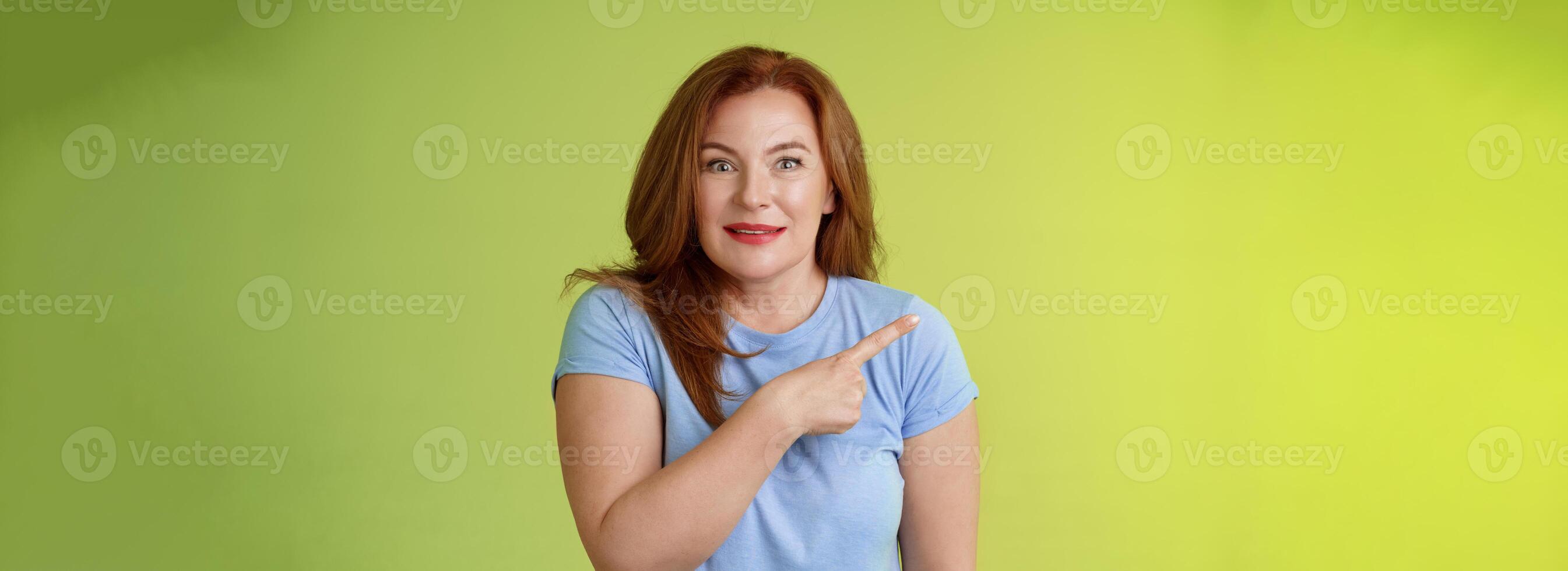 Surprised excited middle-aged wondered redhead woman pointing left amused standing thrilled joyful green background look camera curious interested cannot wait check-out great promo photo
