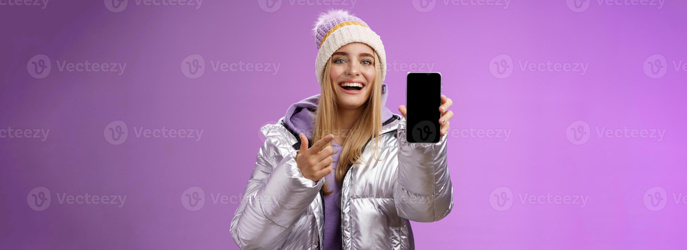 Satisfied amused good-looking blond girl suggest take look smartphone display smiling happily pointing mobile phone delighted talking about awesome new app features, standing purple background photo