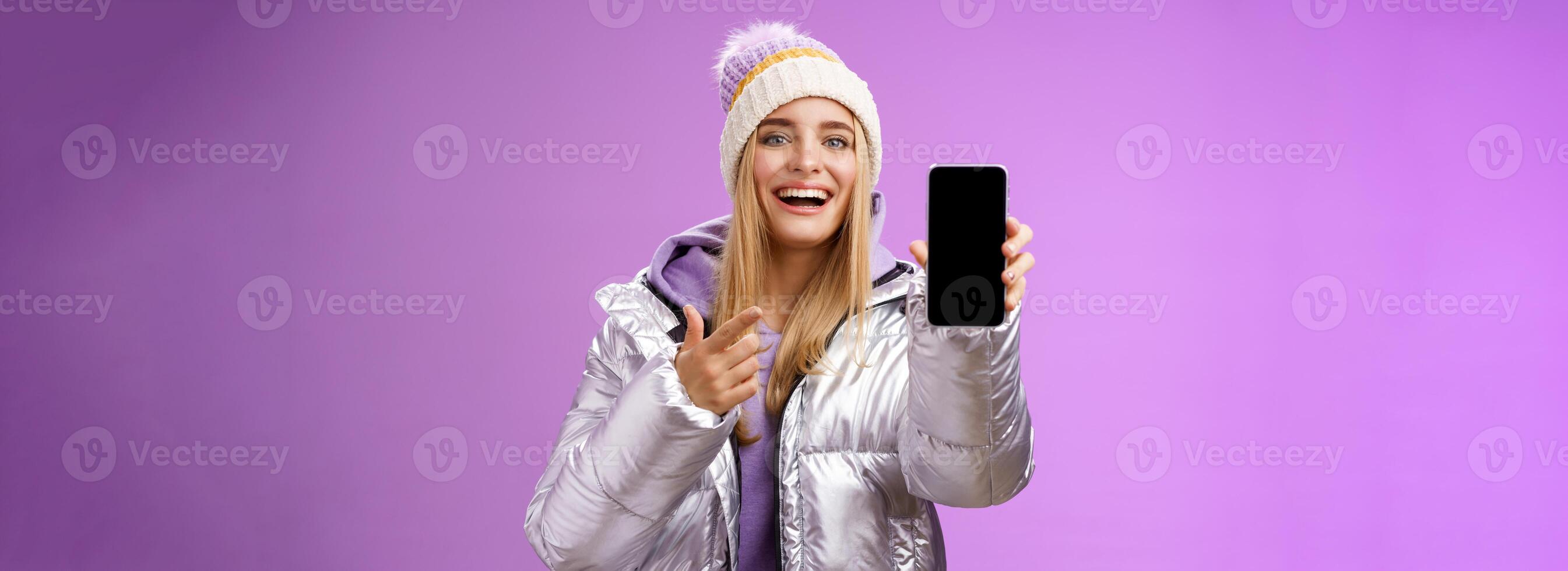 Satisfied amused good-looking blond girl suggest take look smartphone display smiling happily pointing mobile phone delighted talking about awesome new app features, standing purple background photo