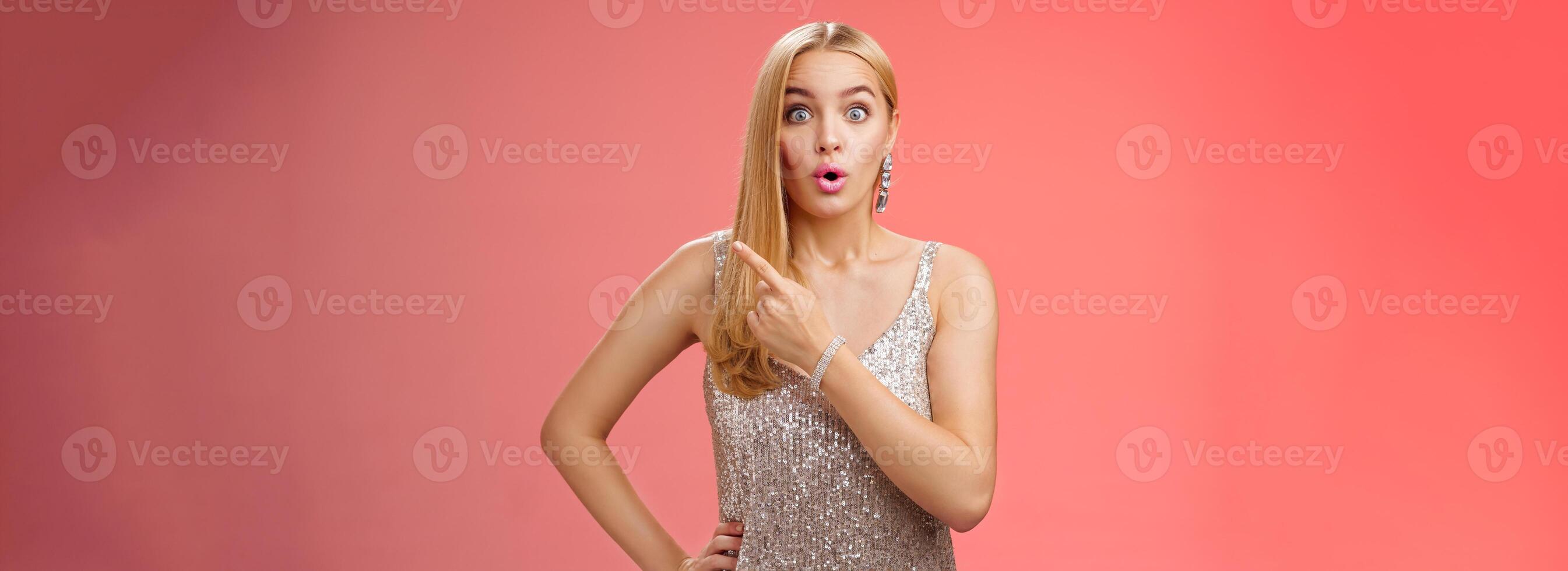 Wondered amused attractive blond woman talking party widen eyes say wow pointing questioned right direction see famous person standing surprised impressed gaze camera, red background photo