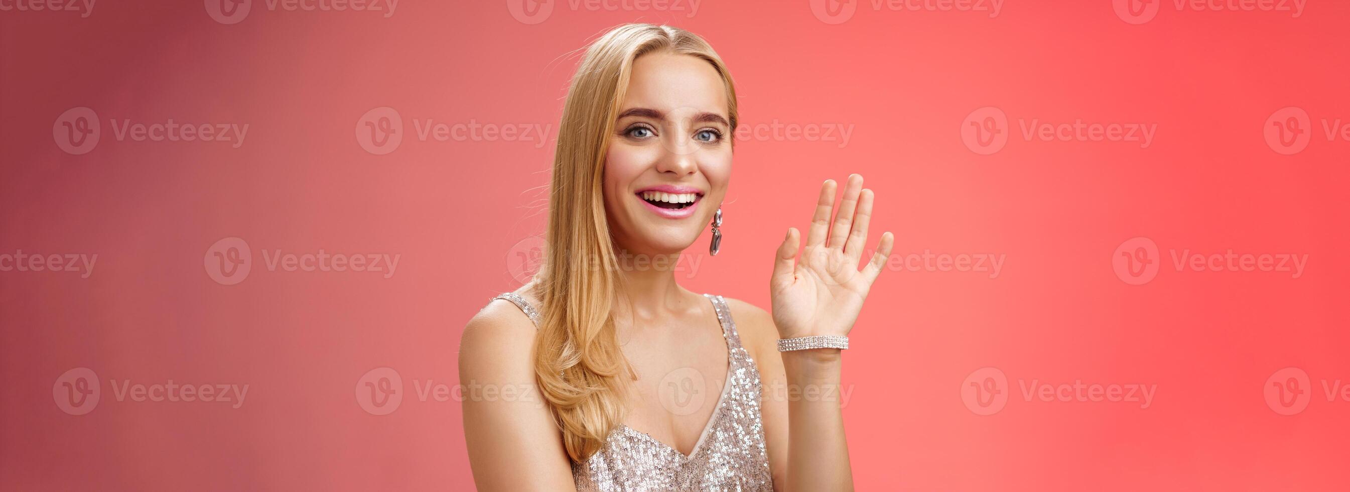 Waist-up studio shot friendly attractive elegant tender blond woman say hi waving raised hand greeting welcoming friend smiling delighted introducing herself hello gesture, red background photo