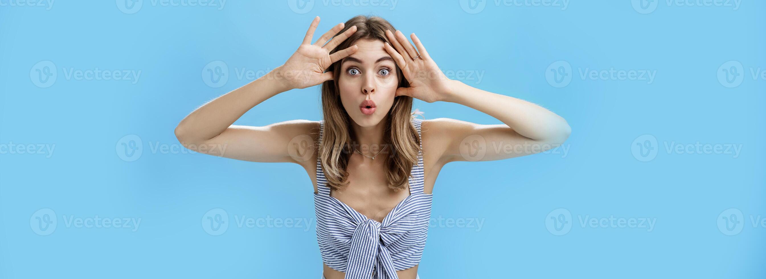 Woman looking surprised and impressed at camera after opening eyes after waiting for surprise. Emotive and playful good-looking european female holding palms near forehead folding lips curiously photo