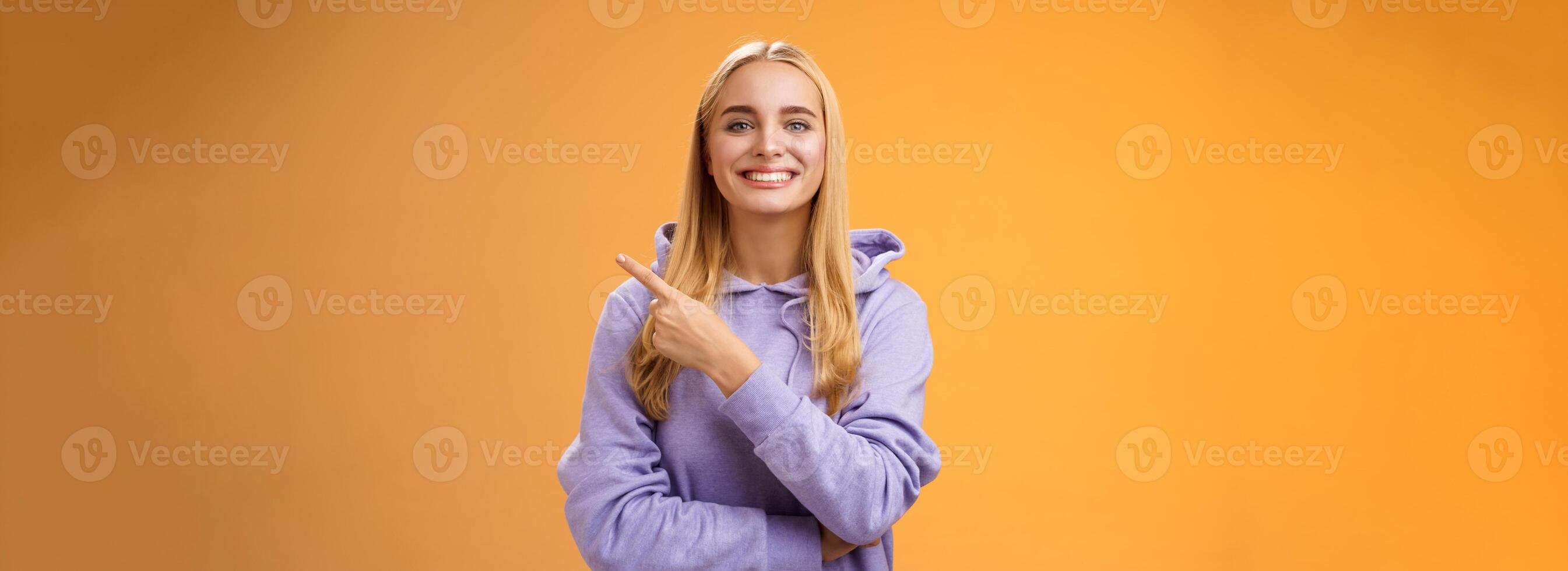 Charismatic modern millennial cute blond woman in hoodie smiling happily discussing cool new promo pointing right recommend check out visit interesting store near corner, orange background photo