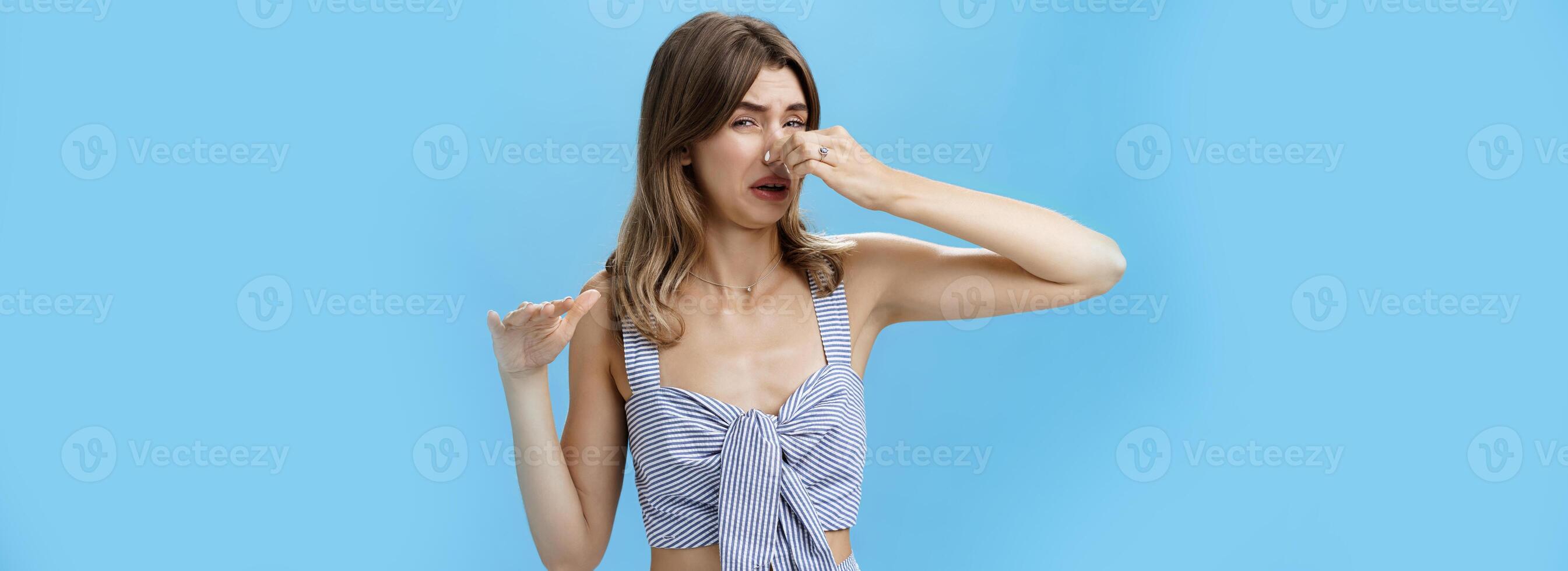 Gross smells awful. Portrait of displeased funny young slim woman with tattoo squeezing nose with two fingers not sniff disfusting reek crying from stink standing intense over blue background photo