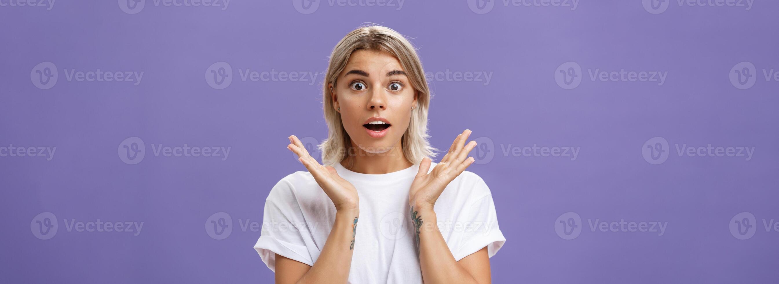 Close-up shot of charmed and impressed good-looking caucasian girlfriend with blond hair and tattoos on arms raising hands in amazed and excited gesture opening mouth and gazing mesmerized at camera photo