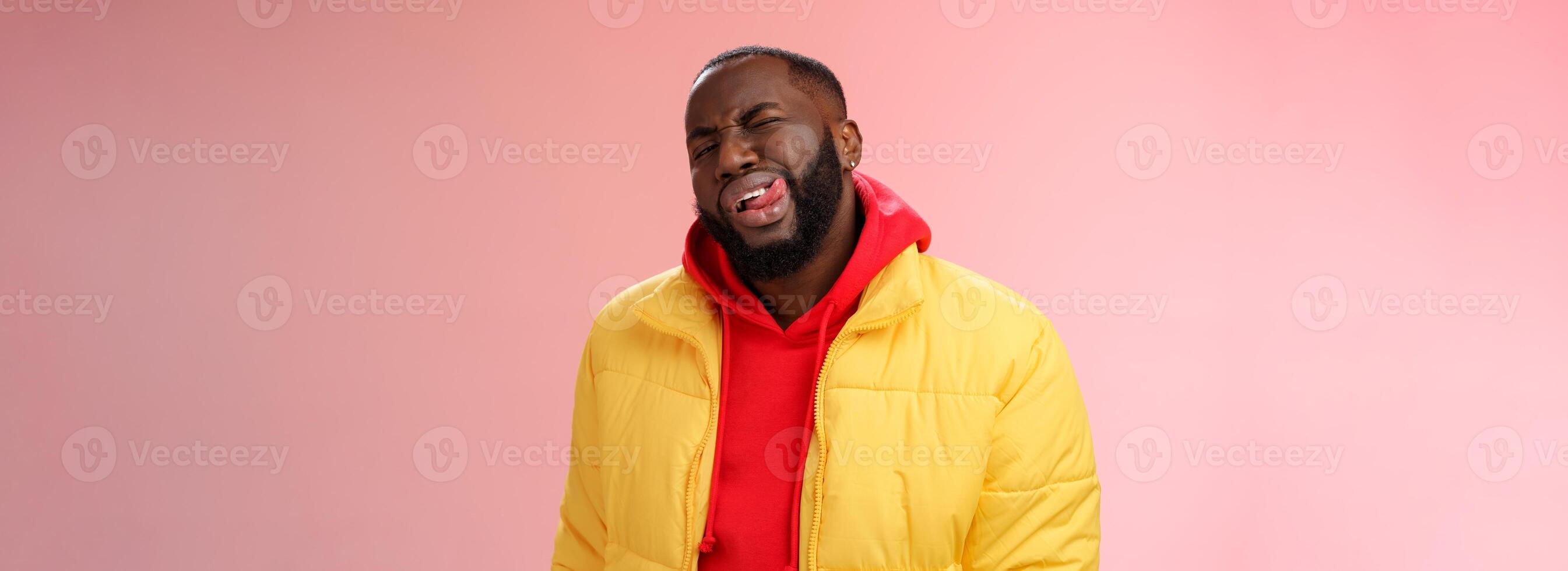 Cheeky stylish funny african bearded man in yellow trendy jacket red hoodie show tongue sassy flirty gaze winking camera trying impress woman pretend macho, standing pink background photo