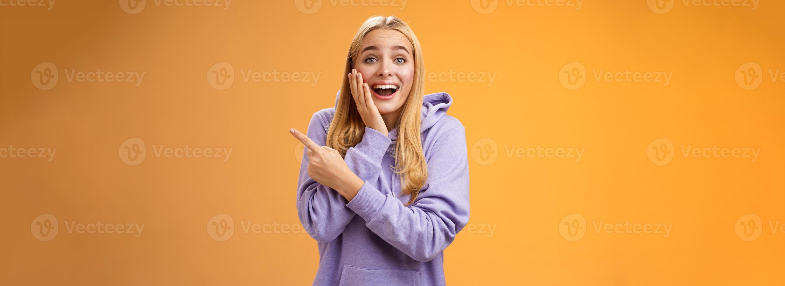 Amused excited speechless happy young blond woman 25s in hoodie cannot believe luck happiness pointing right amazed smiling broadly touch cheek surprised receive awesome unexpected gift photo
