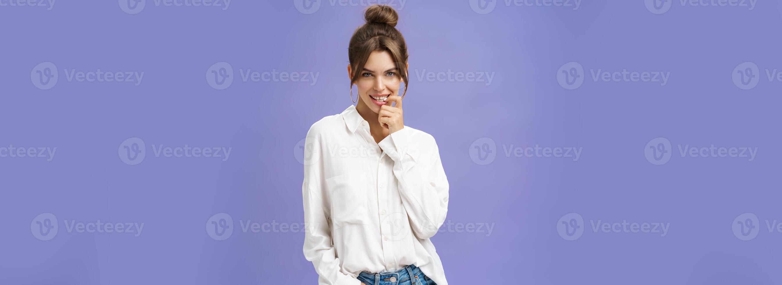 Sensual and flirty self-assured attractive woman in stylish white blouse with good self-esteem holding hand in pocket biting finger showing seducing skills looking daring at camera over purple wall photo