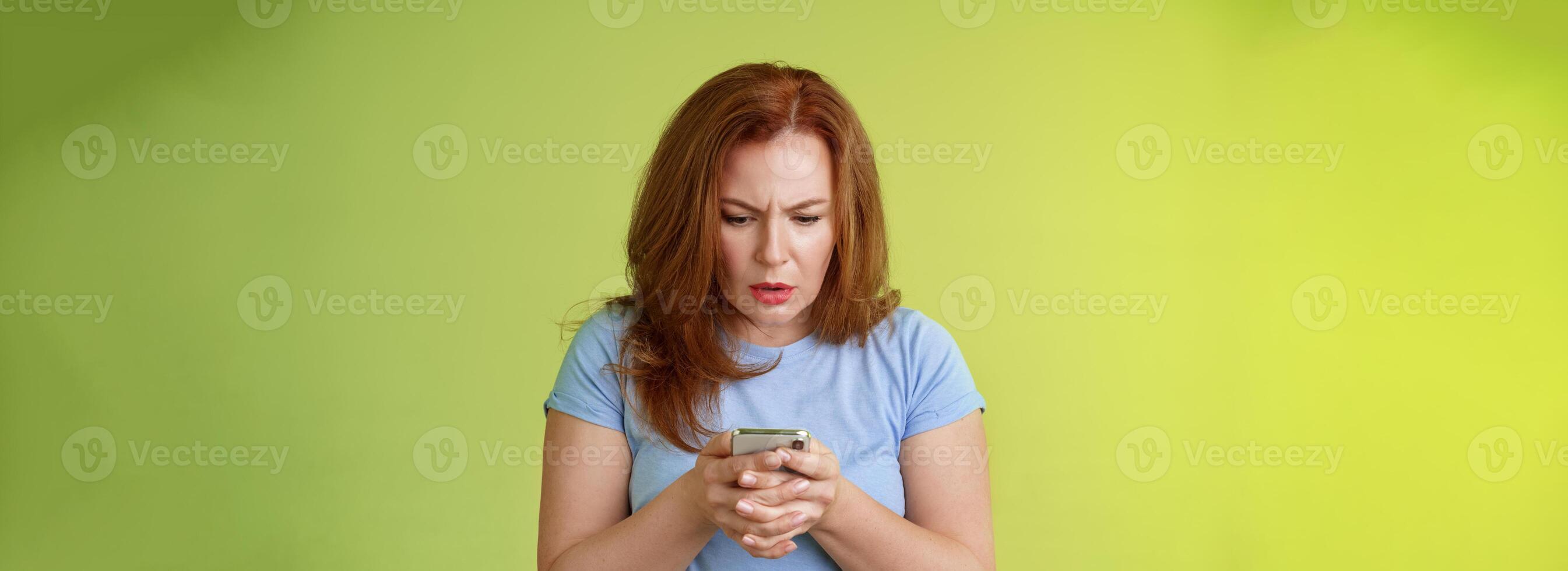 Confused unsure redhead middle-aged woman learn how use social media trying understand emoji look intense focused smartphone display reading important news message stand green background photo
