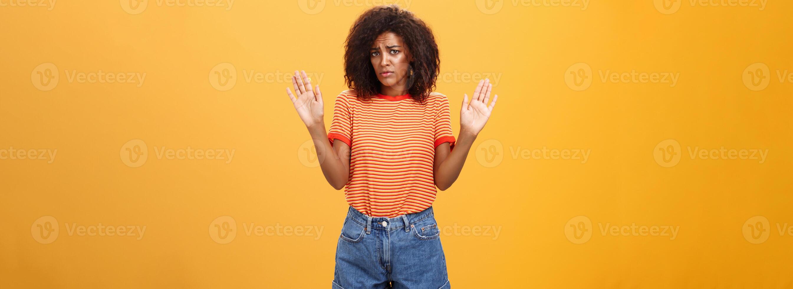 Silly insecure and sad dark-skinned female model in trendy striped t-shirt and shorts raising arms in surrender frowning being uninvolved and unaware standing clueless over orange background photo