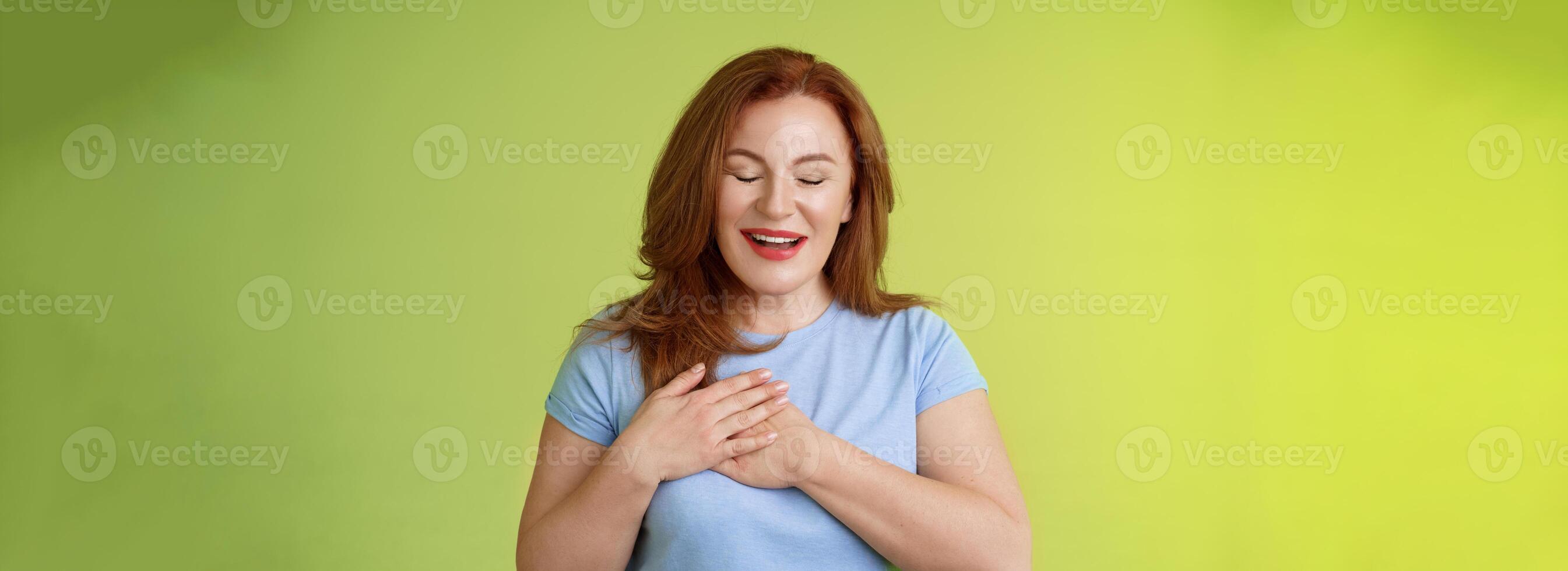 Fascinated cute redhead passionate middle-aged woman sighing lovely touch heart close eyes smiling delighted express admiration temptation feeling appreciation grateful emotions green background photo