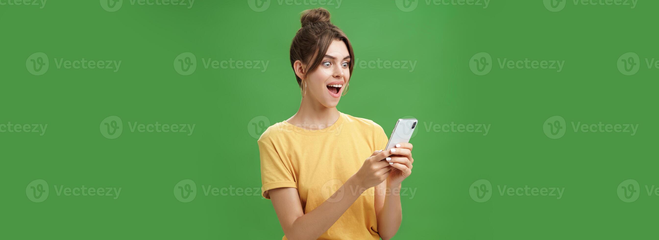 Woman reading surprising satisfying message in smartphone opening mouth from excitement, smiling amazed looking astonished at cellphone screen posing against green background in casual yellow t-shirt photo