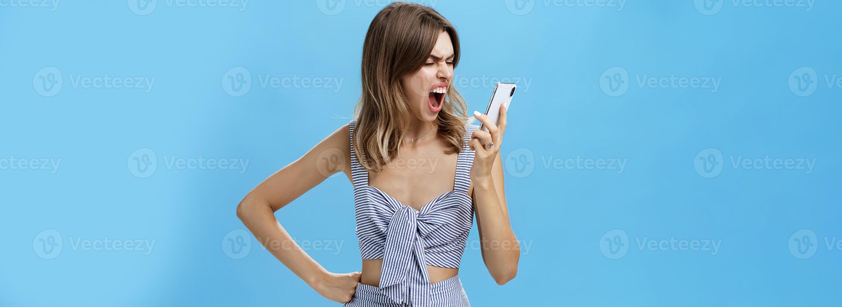Woman yelling at smartphone with sick and tired look being agressive and angry swearing on boyfriend who dumbed her on phone holding hand on waist looking with hate at cellphone screen photo