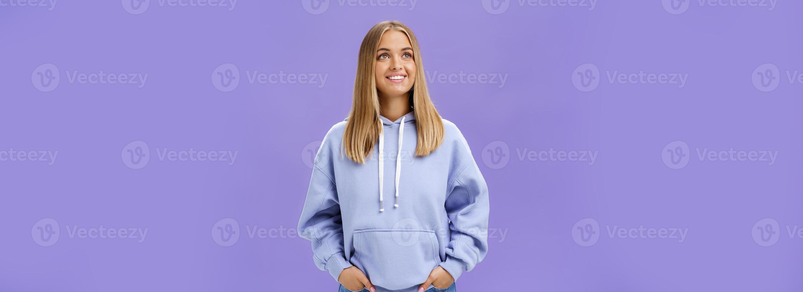 Winter comming. Dreamy kind and cute young girl with straight fair hair and tan wearing hoodie looking at upper left corner with nice white smile and admiring expression holding hand in pockets photo