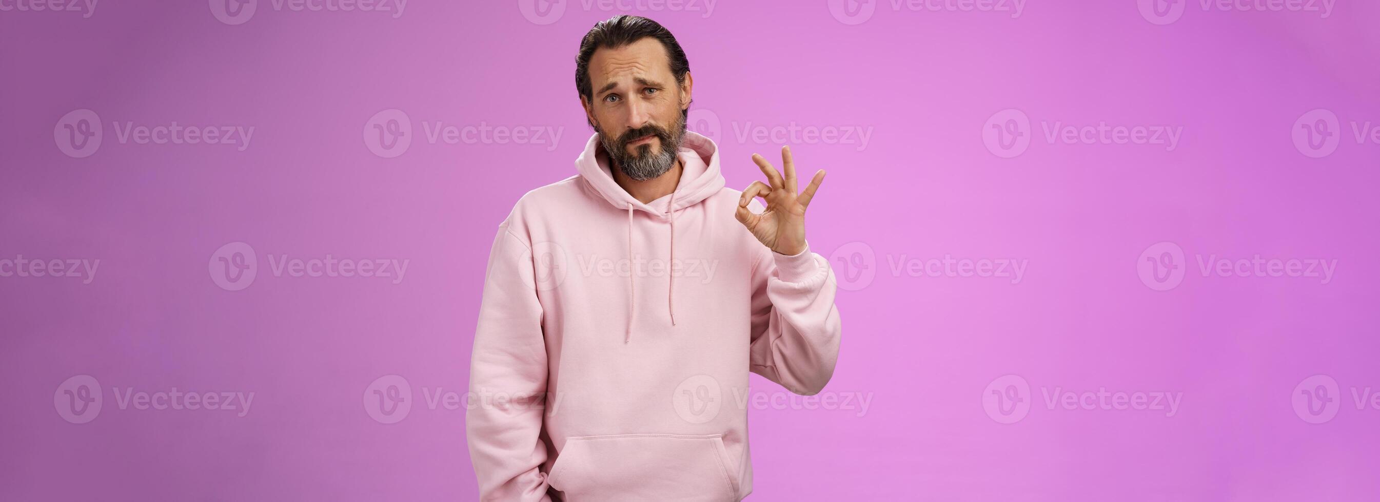 Not bad may better. Portrait handsome mature bearded father wearing trendy hoodie frowning awkward show okay ok normal gesture unimpressed but agree, standing purple background doubtful unsure photo