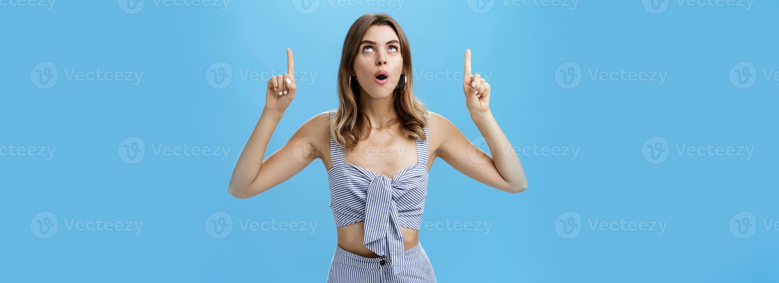 Waist-up shot of curious good-looking stylish modern woman dropping jaw saying wow while lifting head up looking and pointing upwards with amazed, surprised expression posing against blue background photo