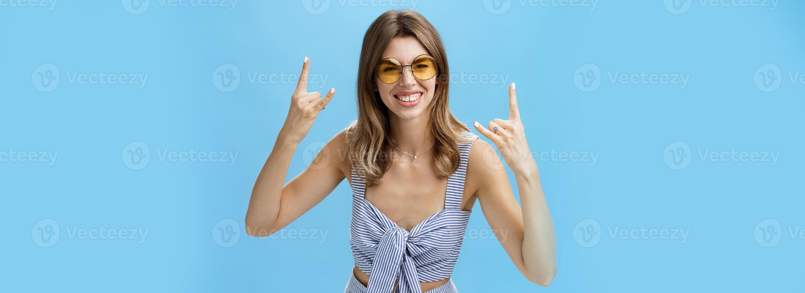 Girl ready to rock-n-roll this party wearing best matching outfit from wardrobe and cool sunglasses showing rock gestures, smiling excited having fun listening music on concert over blue wall photo