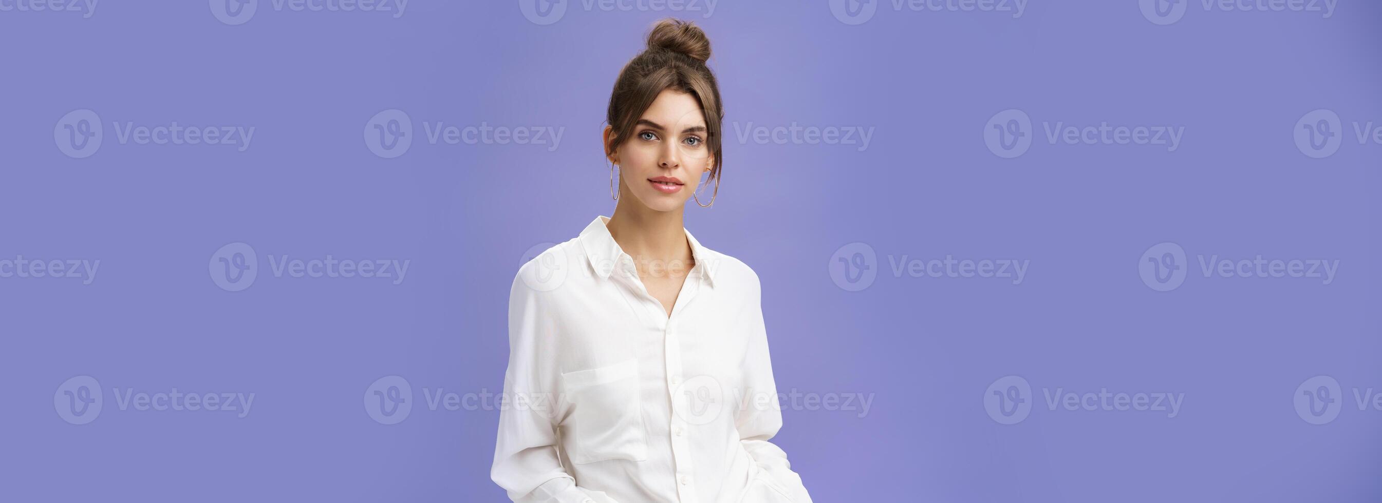 Portrait of good-looking confident female professional in white blouse and jeans holding hands in pockets looking self-assured at camera dressing for office work posing against purple background photo