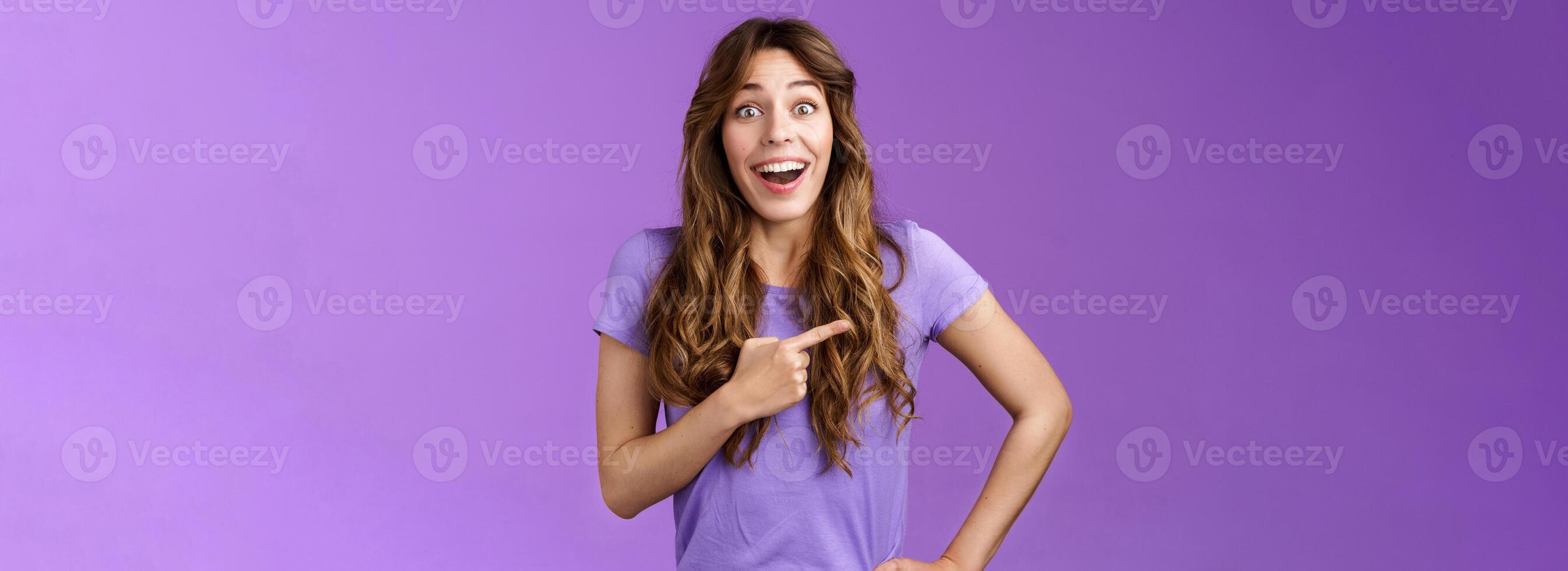 Lively excited enthuasitic good-looking woman curly hairstyle open mouth fascinated look admiration joy share awesome place location pointing left smiling broaldy thrilled purple background photo