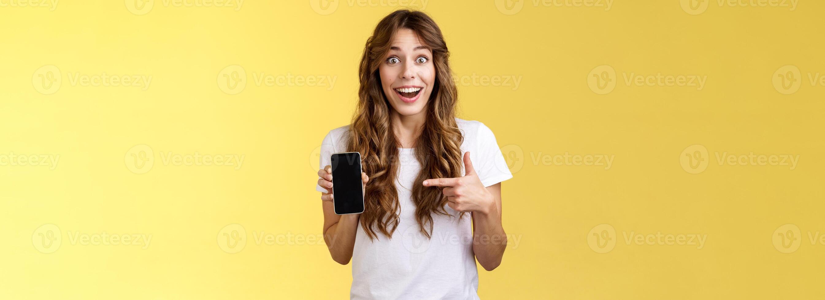 Surprised happy lucky girl winning online internet lottery smiling broadly hold smartphone pointing blank mobile phone screen showing display grinning excited enthusiastic stand yellow background photo