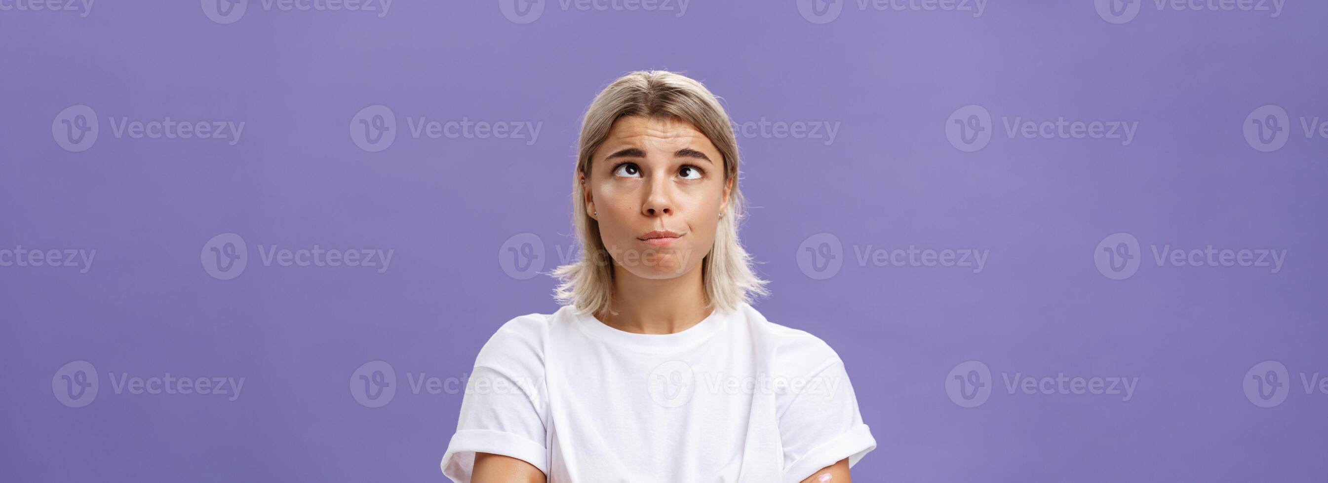 Mind going crazy. Portrait of funny tired attractive young woman with fair hair and tanned skin squinting rolling eyes aside and looking up fooling around over purple background from boredom photo