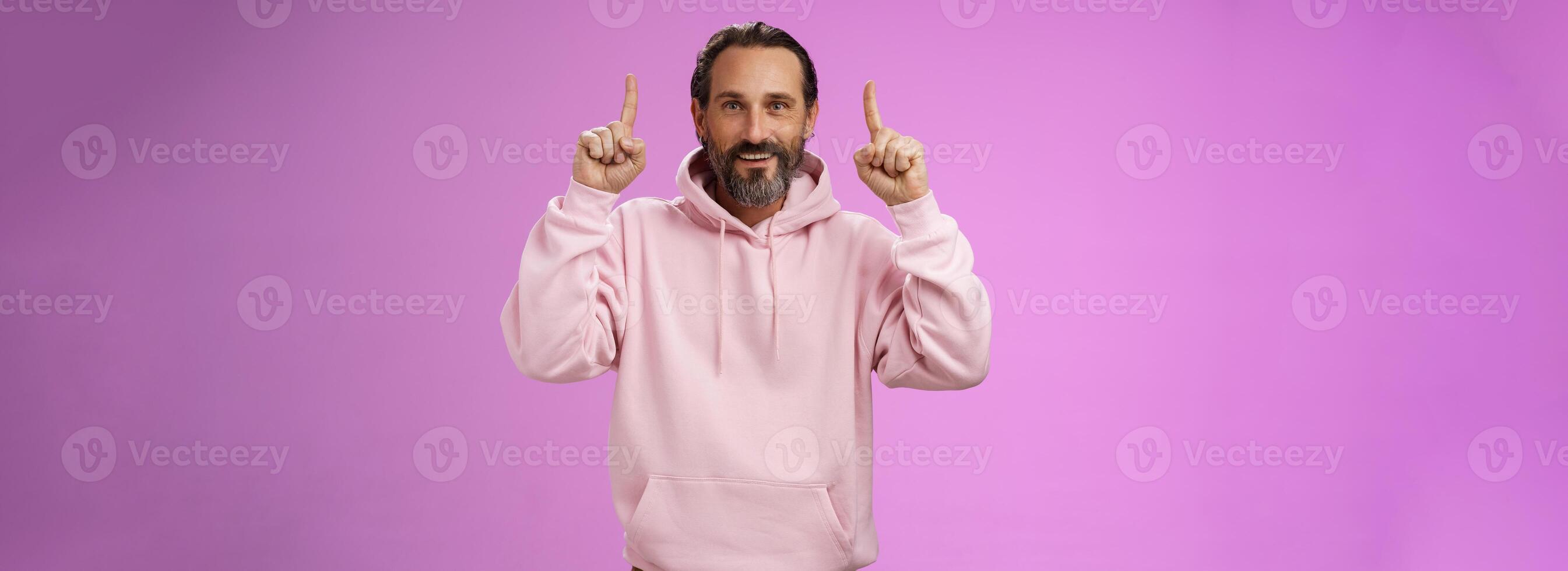 Handsome mature 50s man grey hair beard wearing stylish hipster loose hoodie pointing up index fingers showing awesome place hang out unwilling grow old standing positive lucky purple background photo