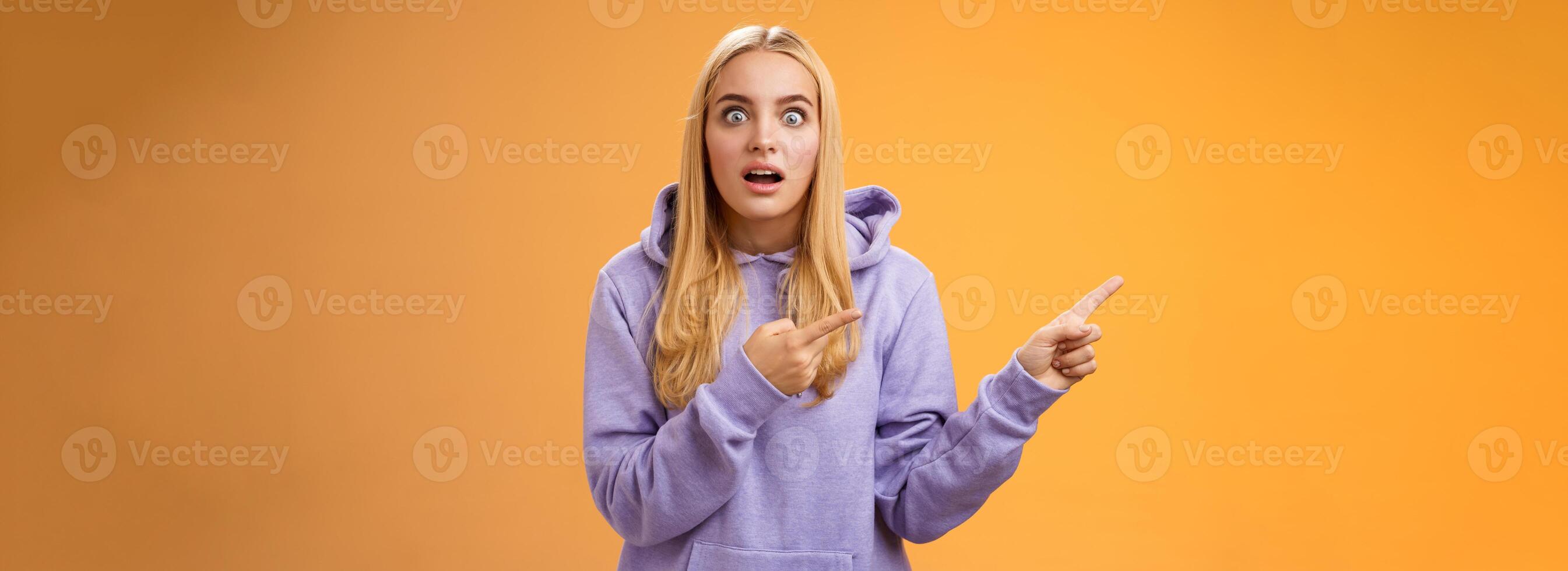 Wow astonished impressed good-looking blond european female in hoodie pop eyes wondered surprised questioned drop jaw pointing left excited asking if gossip true, standing orange background photo