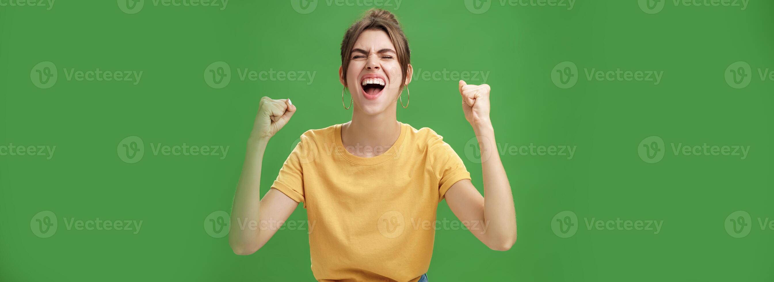 Waist-up shot of charismatic energized and excited female in yellow t-shirt closing eyes yelling from joy and happiness raising hands in cheer celebrating successful news over green background photo