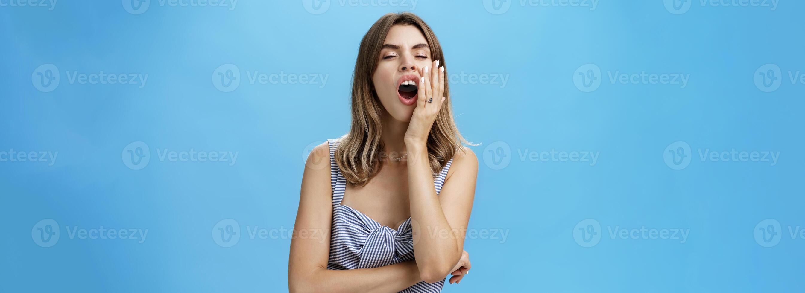 Charming stylish and attractive female manager in matching outfit with cute gapped teeth yawning covering opened mouth with palm gazing sleepy and indifferent at camera feeling bored over blue wall photo
