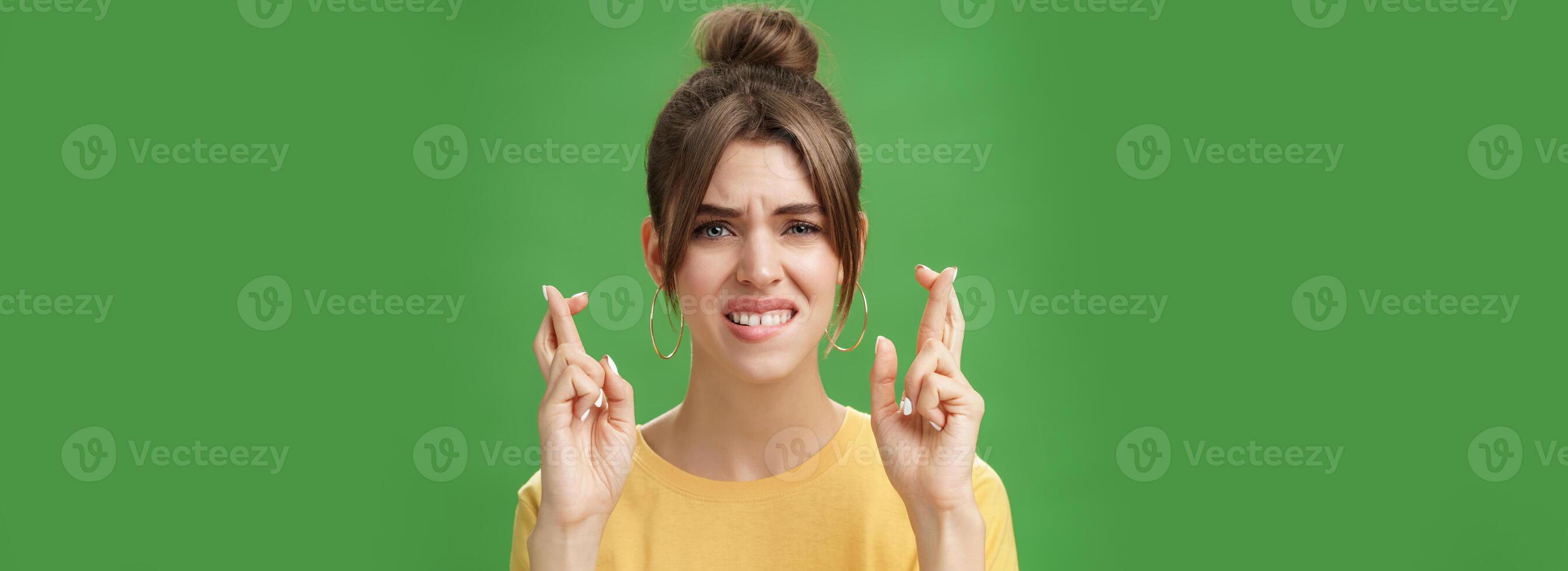 Close-up shot of nervous woman praying for having positive answer crossing fingers for good luck biting lower lip anxiously and frowning being worried while hoping to get job over green background photo