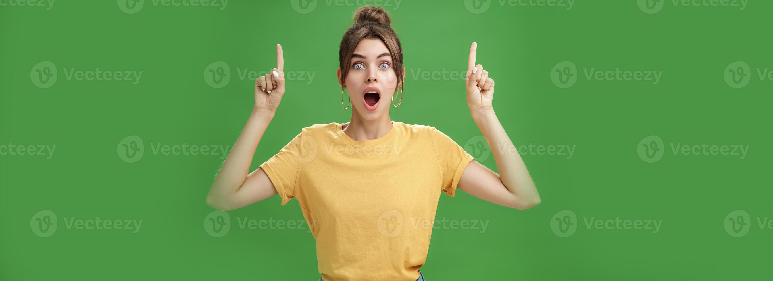 Waist-up shot of interested and surprised amazed good-looking female in yellow t-shirt with tattoo dropping jaw impressed and astonished raising hands pointing up questioned against green background photo