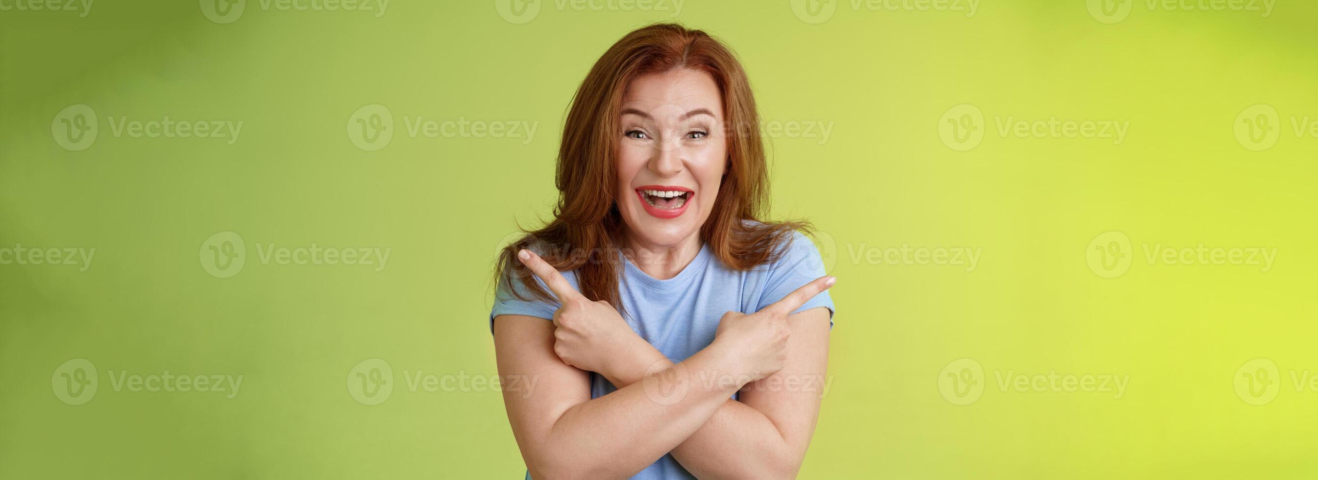 Happy carefree entushiastic redhead funny mature female having fun positive attitude cross arms body pointing sideways show left right products laughing happily like both choices green background photo