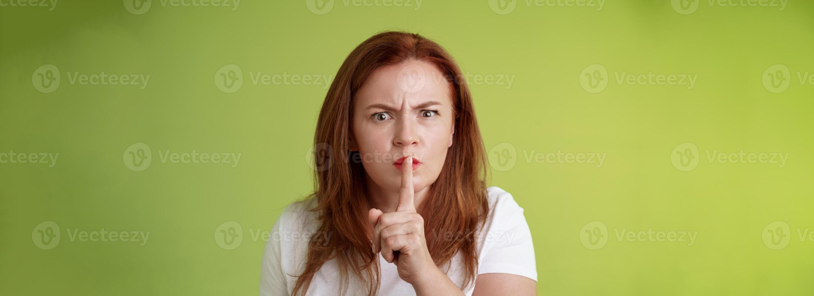 Not speak during exam. Strict serious-looking displeased middle-aged redhead woman frowning disappointed hushing say shush index finger pressed lips keep quiet gesture green background photo