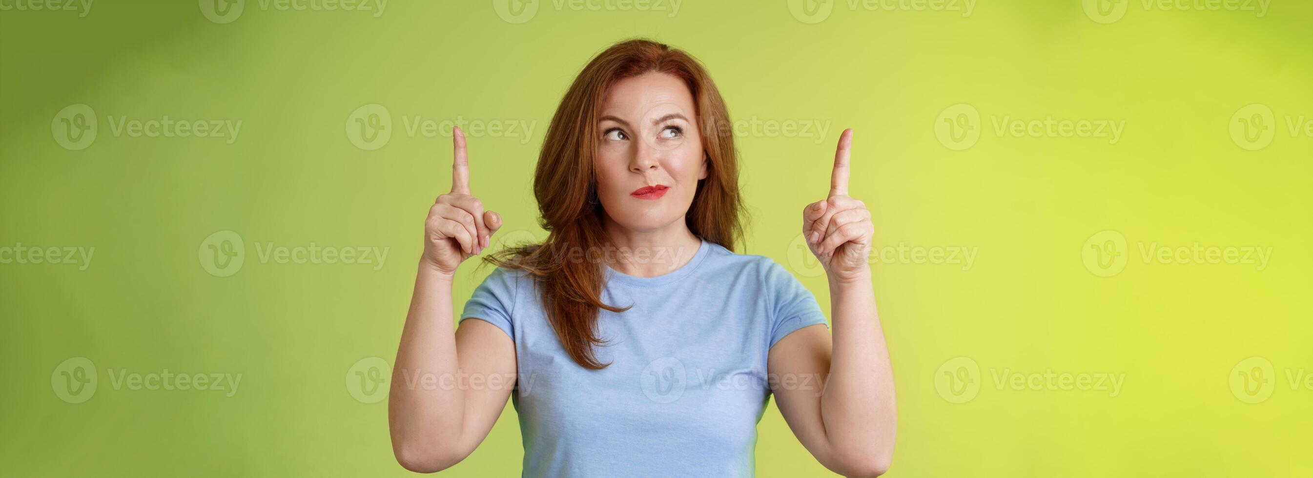 Curious thoughtful cute redhead female middle-aged ginger mother smirking thinking look aside pointing up index fingers deciding choice pondering choosing between variants green background photo