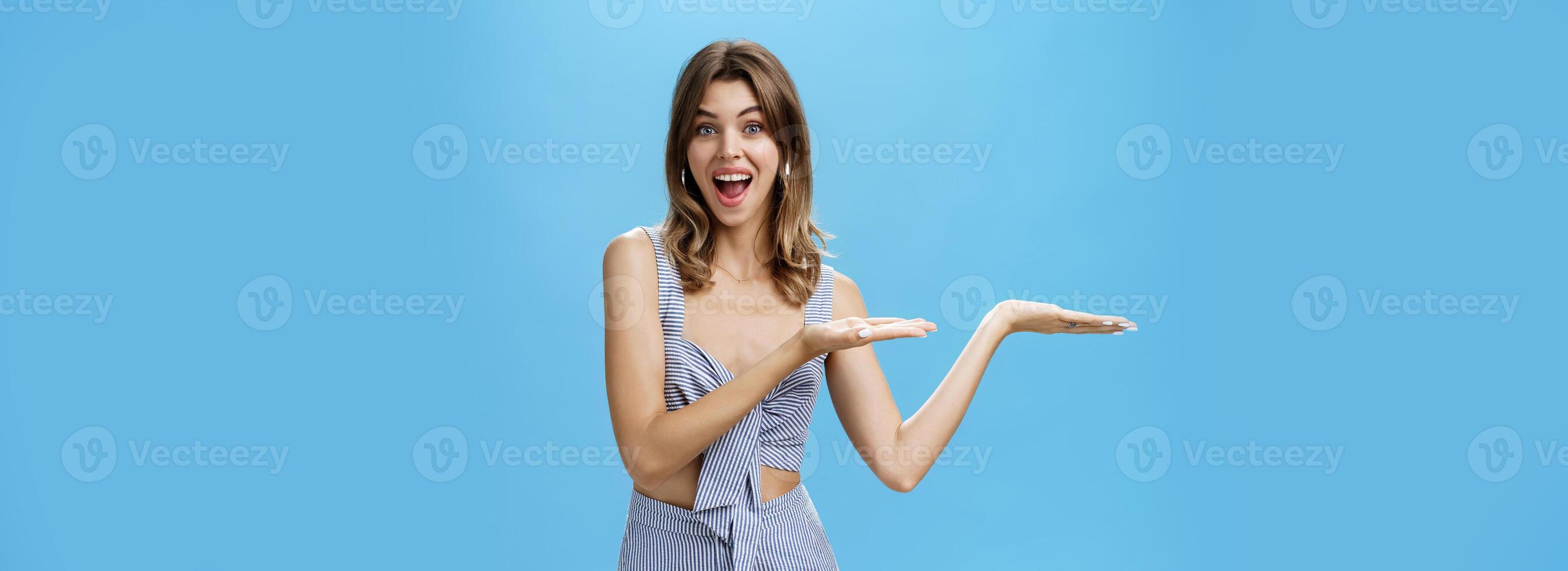 Woman presenting product over copy space. Good-looking charismatic adult tall woman with chestnut hair pointing at left side of wall and smiling broadly as if advertising against blue background photo