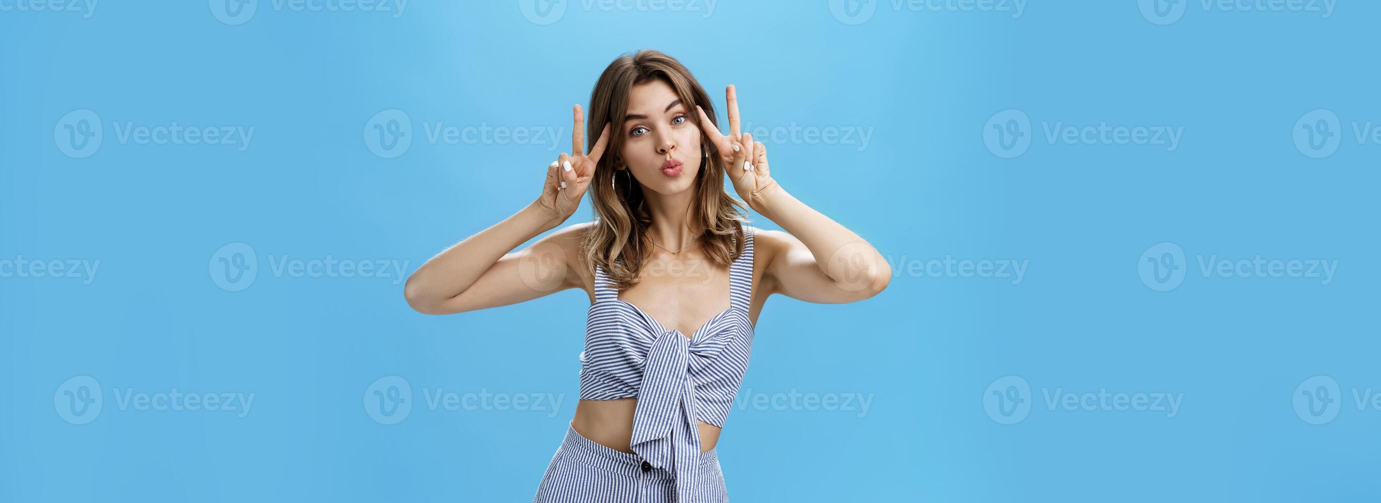 Charming cute unretouched woman with chestnut hair and tattoo showing peace gestures near face folding lips in mwah making silly face while posing over blue background confident and carefree photo
