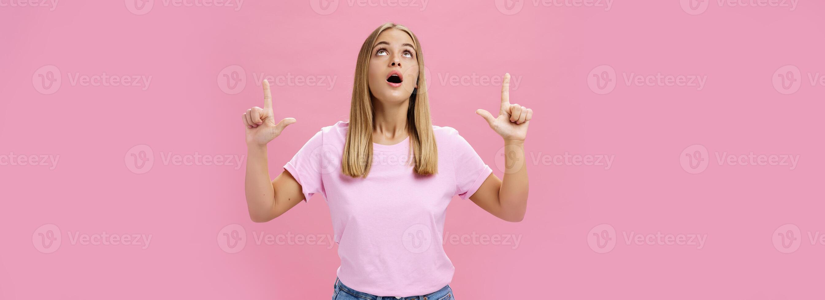 Portrait of amused and impressed curious attractive woman in t-shirt dropping jaw looking and pointing up astonished and intrigued watching interesting object in sky posing against pink background photo