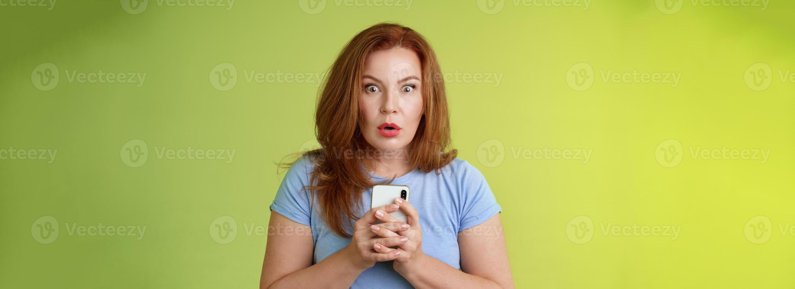 Shocked speechless impressed redhead european middle-aged woman fold lips astonished wow stare camera fascinated react amazed wondered received message hold smartphone intense green background photo
