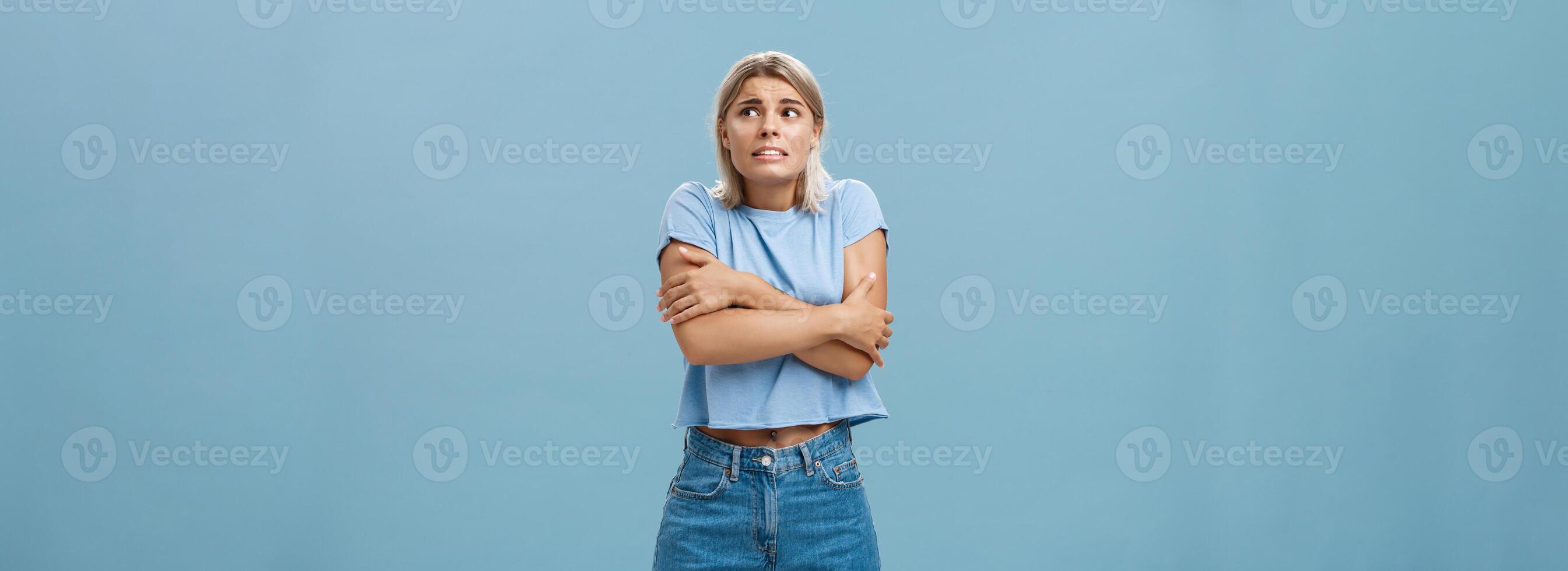 Turn off air-cooler I am freezing in summer. Displeased cute caucasian blonde girl in trendy t-shirt hugging herself while trembling from cold shaking gazing right with worried look over blue wall photo
