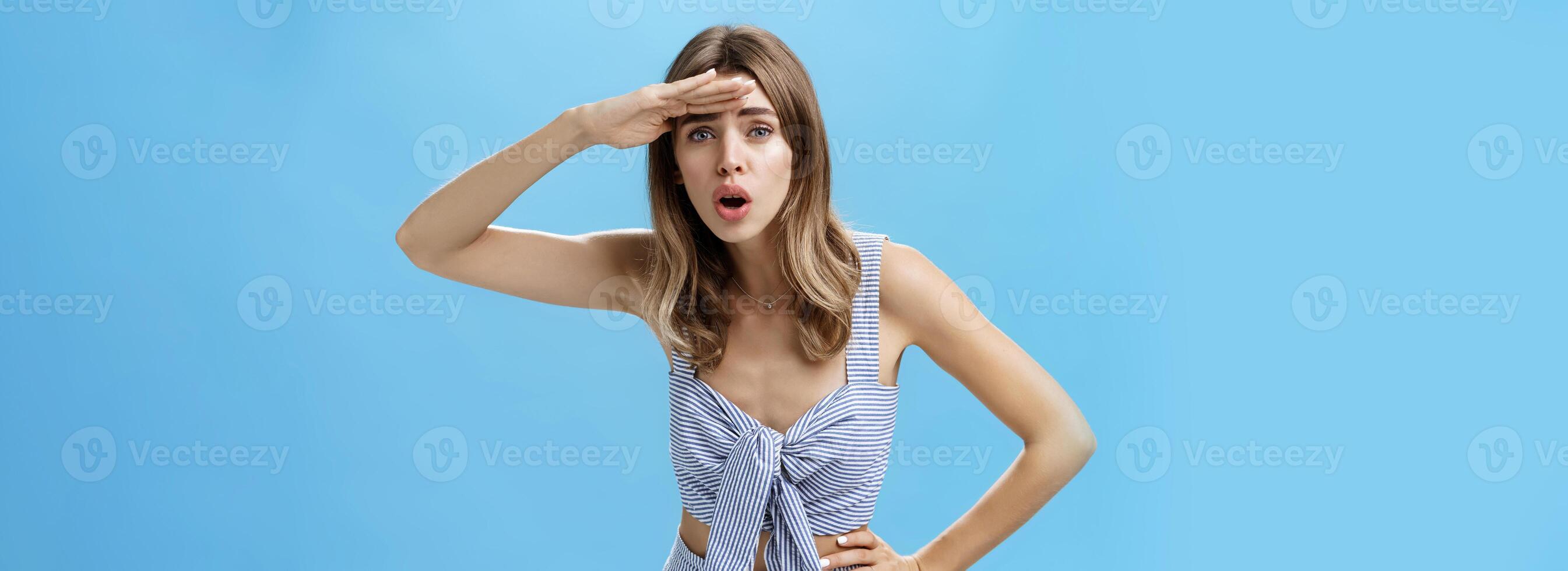 Woman trying see on distance. Portrait of questioned attractive caucasian woman bending holding palm on forehead to protect from sunlight open mouth squinting looking far away searching someone photo