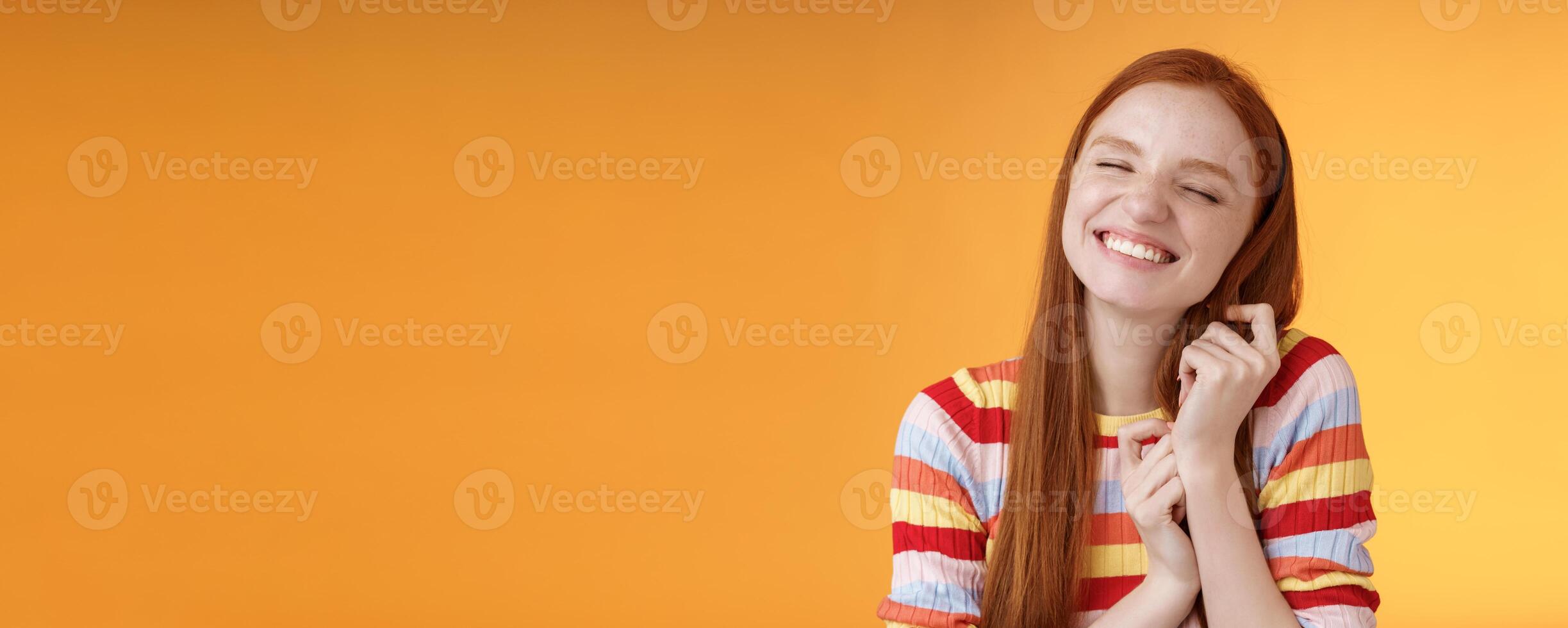 Happy dreamy romantic young tender ginger girl fantasizing creating love story imagination smiling broadly delighted close eyes touching hair strands recalling nice memory, standing orange background photo