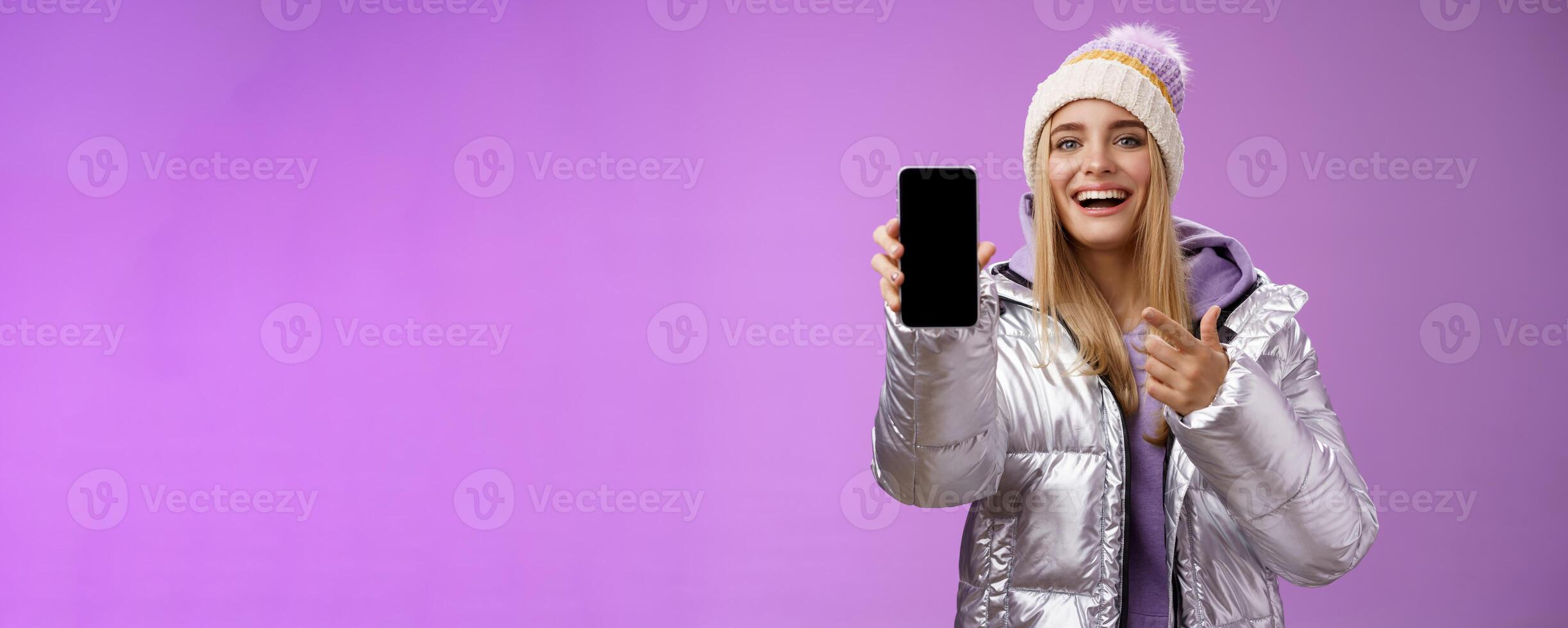 Satisfied amused good-looking blond girl suggest take look smartphone display smiling happily pointing mobile phone delighted talking about awesome new app features, standing purple background photo