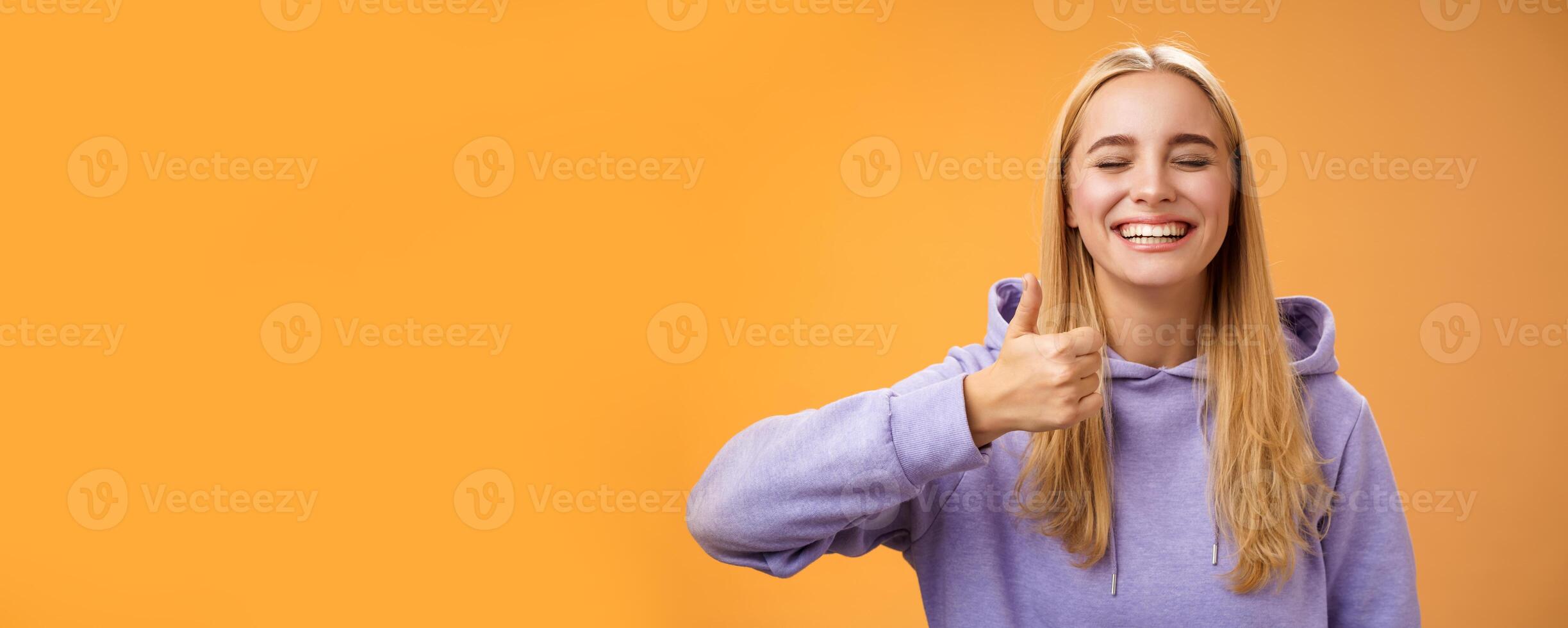 Amused joyful optimistic young excited blond european female model in hoodie smiling broadly close eyes having fun show approval gesture thumbs-up like awesome trip vacation idea, orange background photo