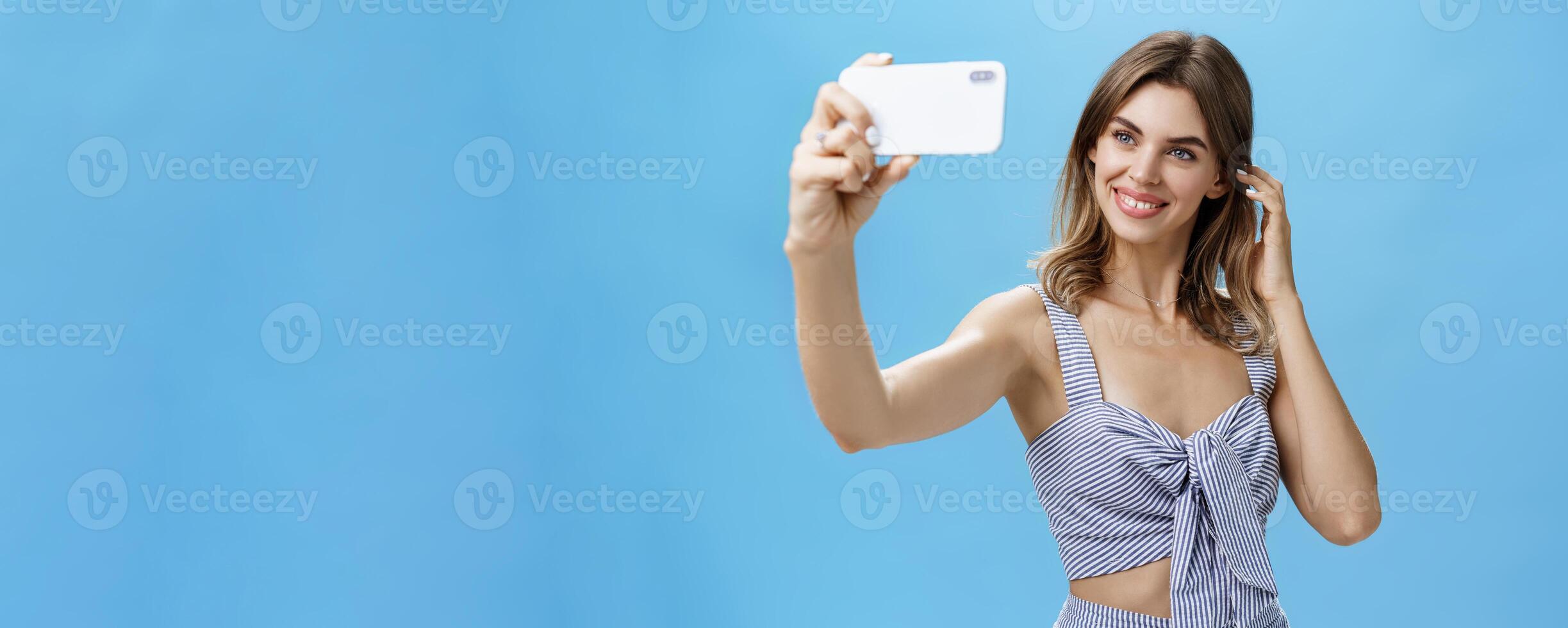 Attractive woman with good self-esteem in stylish matching outfit flicking hair behind ear smiling joyfully at smartphone screen, taking selfie to post in internet and gain followers over blue wall photo