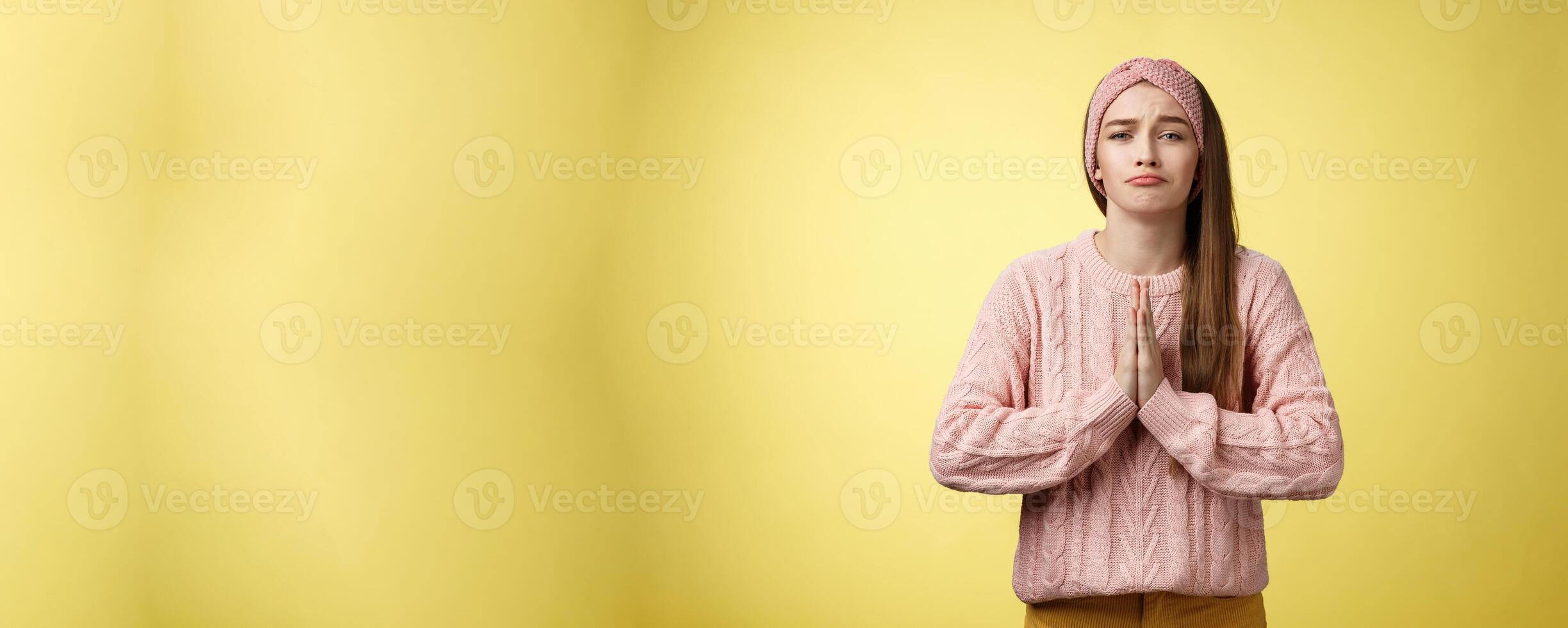 Deliberately sad girl in sweater pressing palms together in pray pouting praying for help, asking favor, begging friend lend cute dress for date, smiling silly, want something over yellow background photo