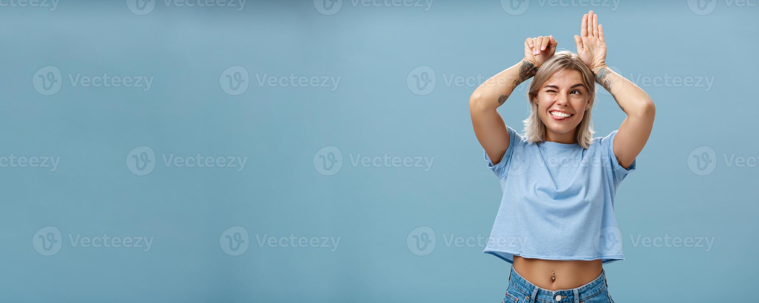 Studio shot of entertained carefree and emotive happy charming woman with tattoos on arms acting like bunny with palms on head winking joyfully smiling and sticking out tongue over blue wall photo