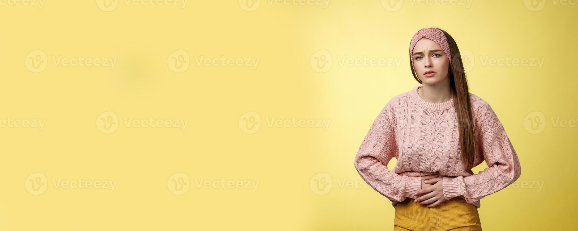 Girl suffering pain in stomach, having painful periods, feeling unwell touching belly grimacing displeased and troubled, having cramps, stooping upset of stomachahe against yellow background photo