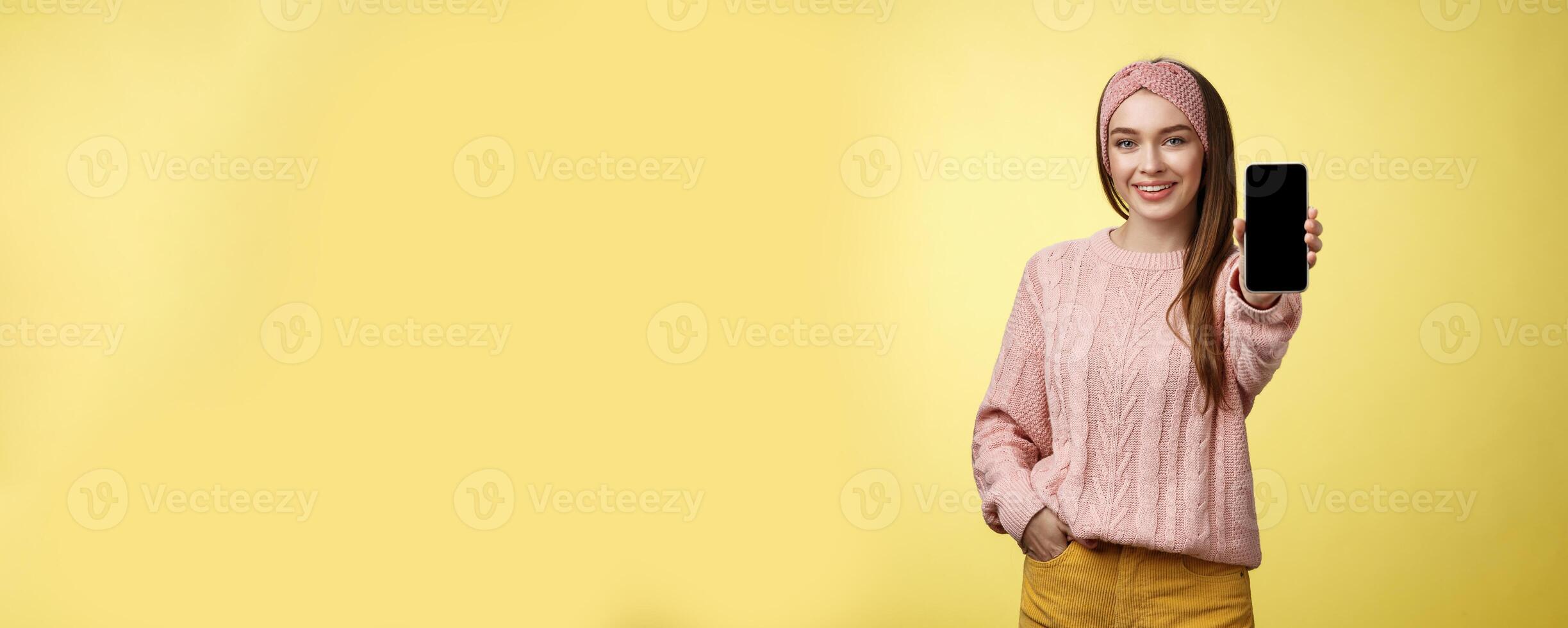 Best smartphone here you go. Charming outgoing young pretty woman in knitted sweater, headband extending hand with phone showing gadget screen smiling recommending cellphone over yellow wall photo