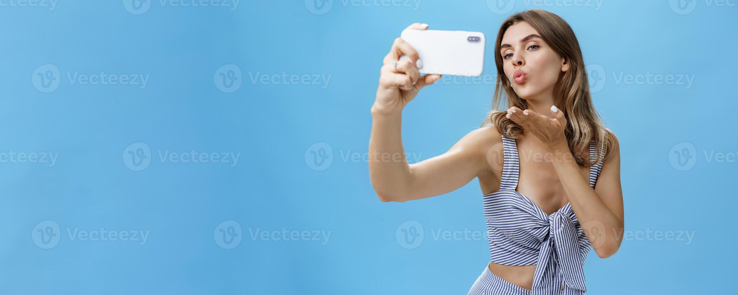 Outgoing and self-assured glamorous woman like taking pictures of herself holding smartphone making sensual and flirty selfie folding lips raising palm to send wind kiss at screen loving followers photo